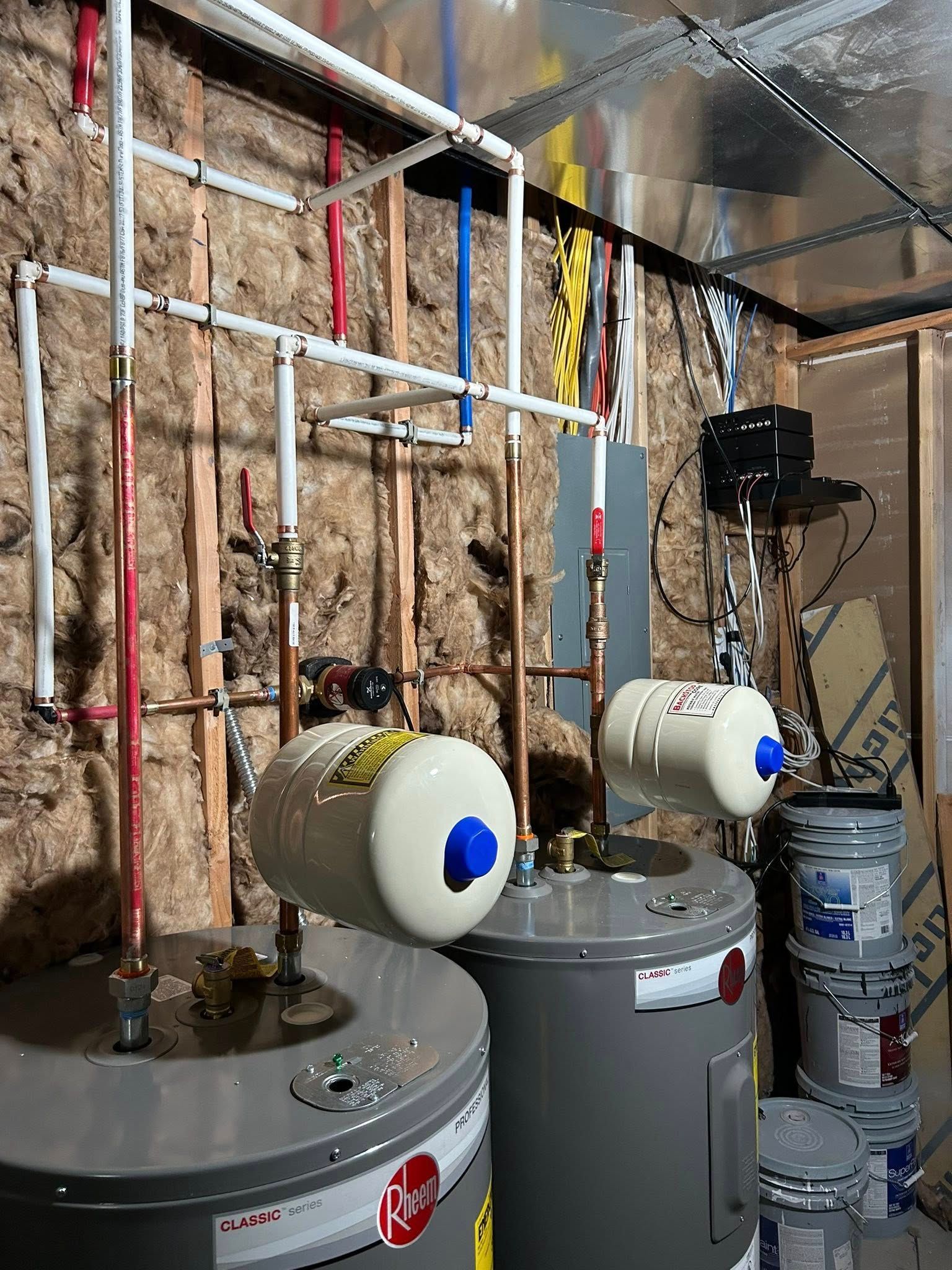 Two water heaters with connected plumbing, insulation, and paint cans in a utility room.