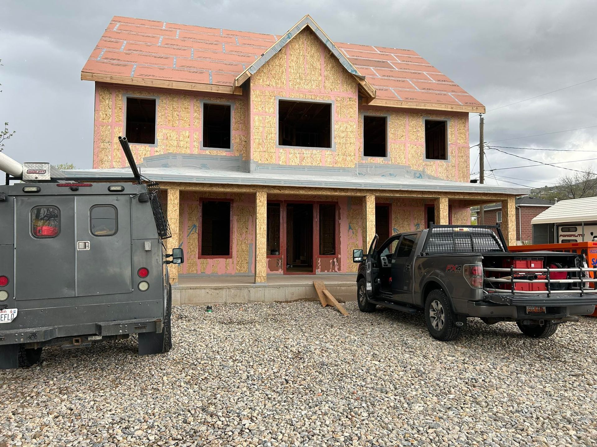 Unfinished two-story house with a truck and armored vehicle parked in front, gravel driveway.