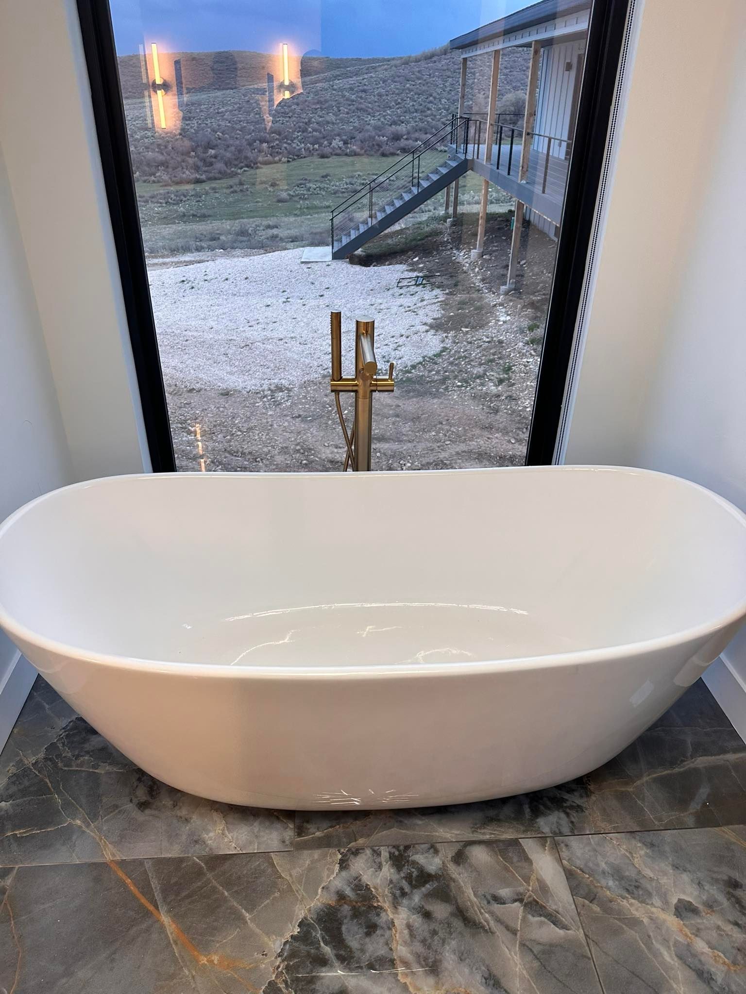 Oval bathtub with gold faucet, set before a large window overlooking a landscape and wooden structure.