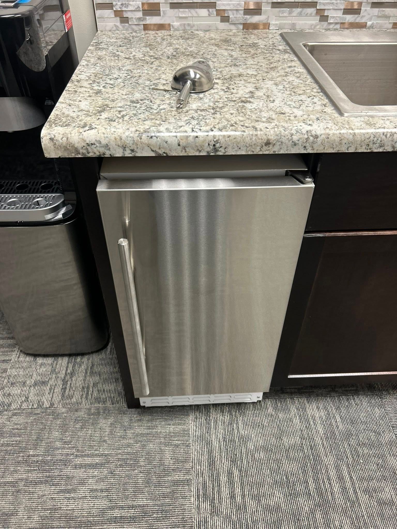 Stainless steel mini-dishwasher in a kitchenette, next to a trash can and sink, under a granite countertop.