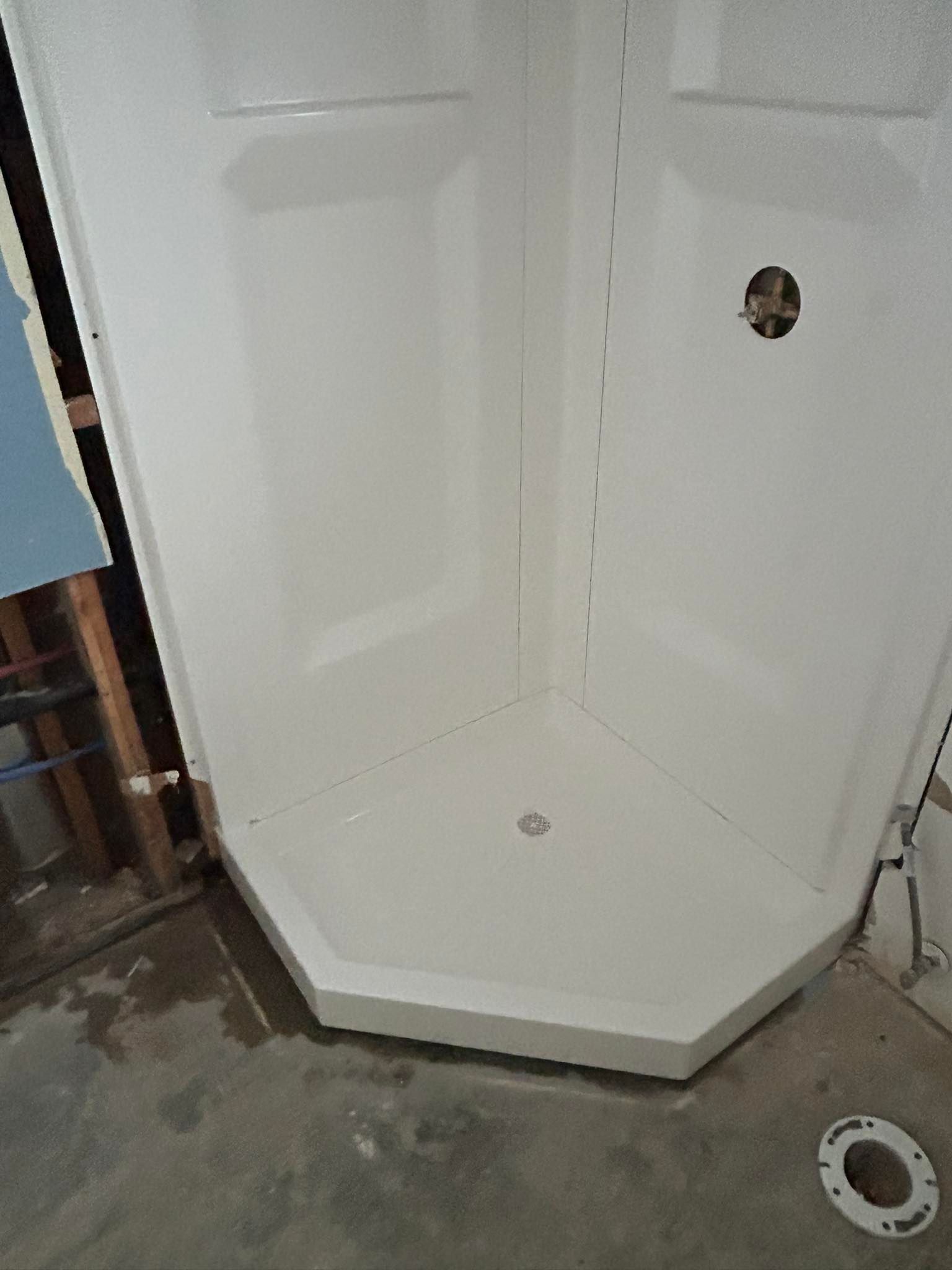 White corner shower stall with a drain and a plumbing access hole, inside a construction site.