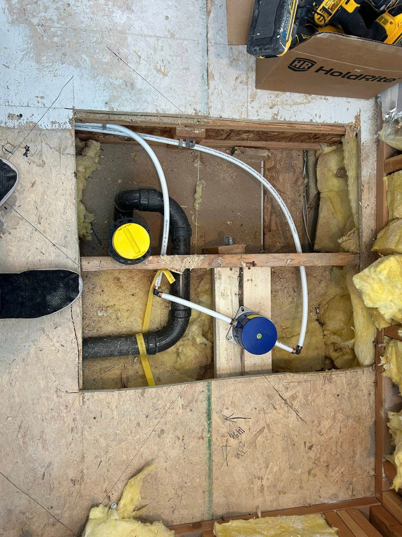 Plumbing repair in a floor opening: Black pipes, yellow and blue components, white tubing, insulation, and wood.
