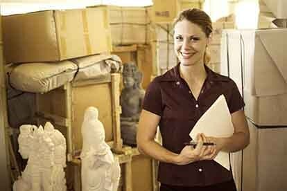 Woman in storage unit, smiling, holding file, surrounded by boxes and sculptures.