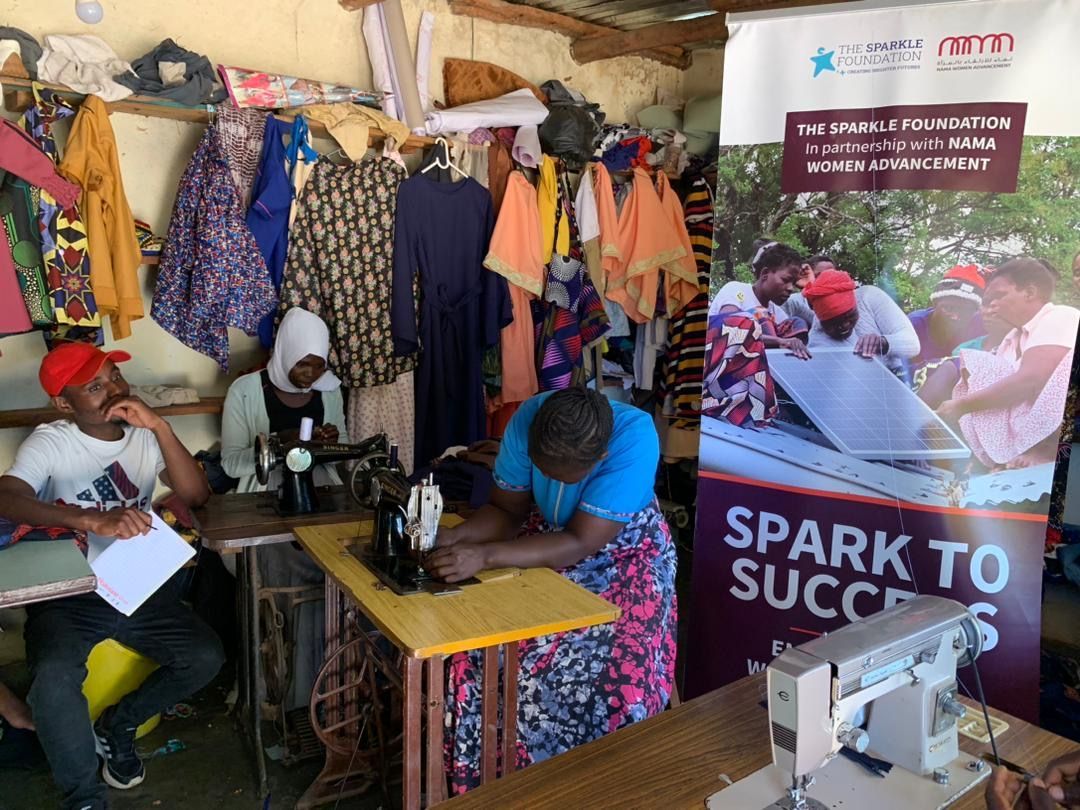 A tailoring trainee operates a sewing machine during a practical assessment at the Spark to Success tailoring training centre in Sakata, Zomba District. Garments made by participants hang around the workshop, while a Spark to Success banner highlights the partnership between The Sparkle Foundation and NAMA Women Advancement.