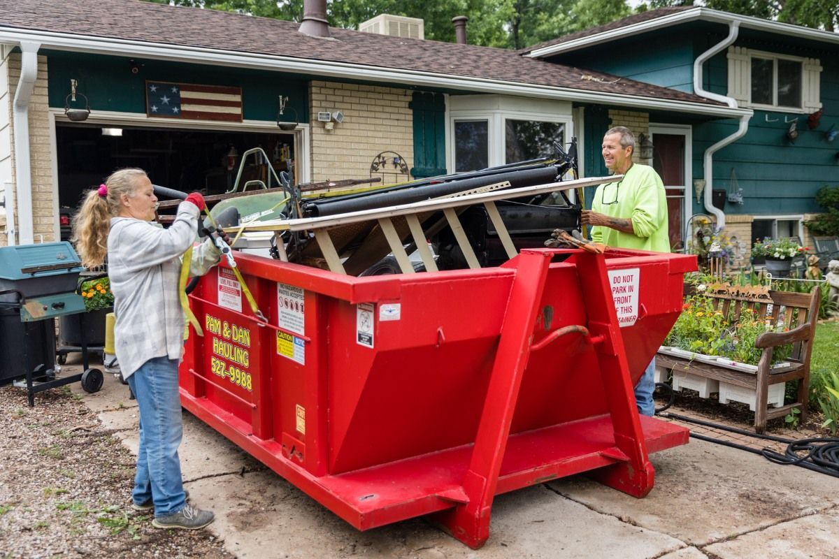 Two people are loading a red dumpster into a driveway in front of a house.