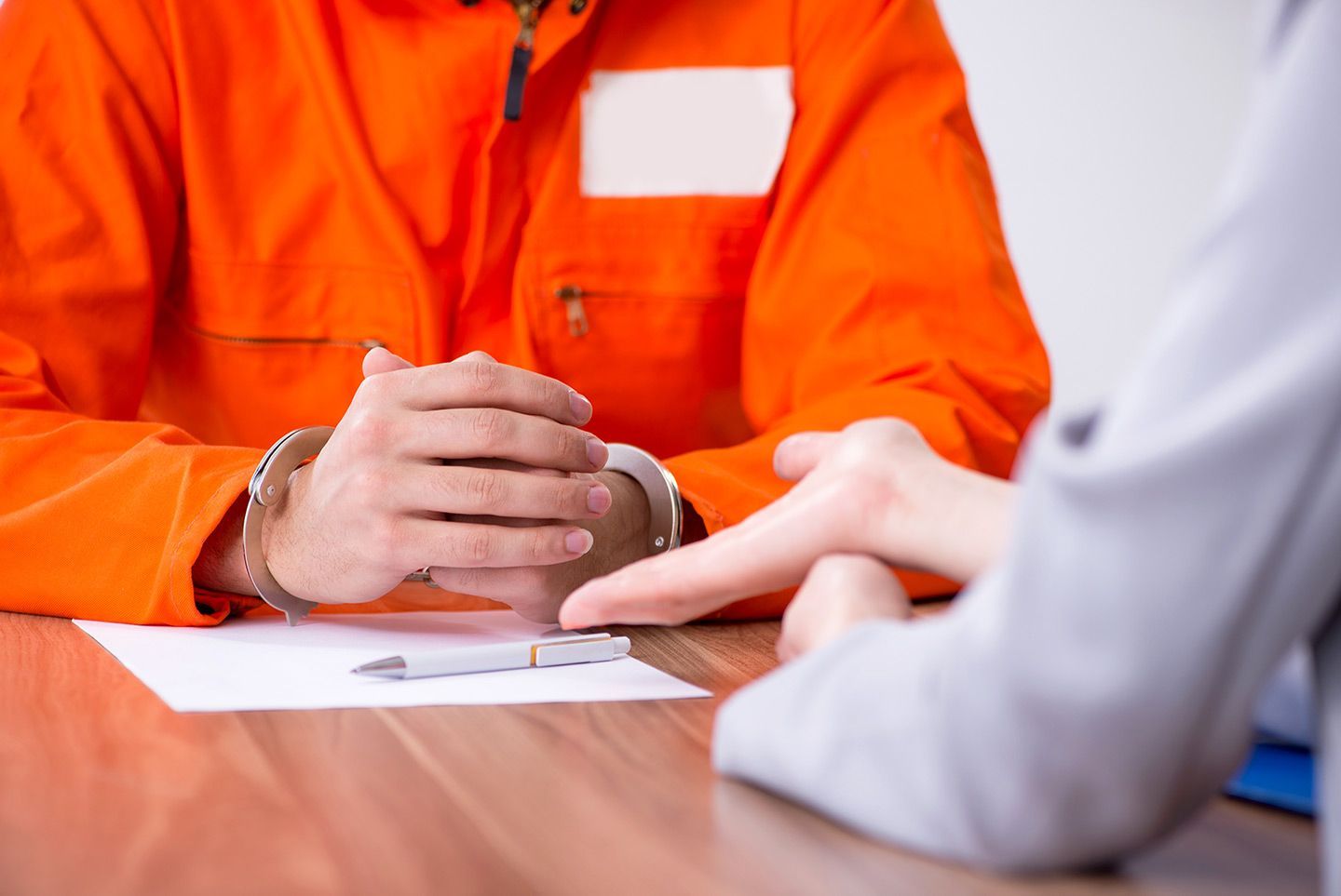 Person in orange jumpsuit with handcuffs, meeting with a person pointing at a paper on the table.