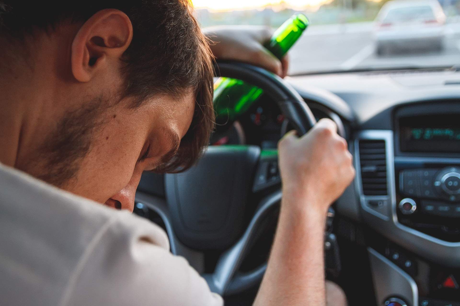 A man is driving a car while holding a bottle of beer.
