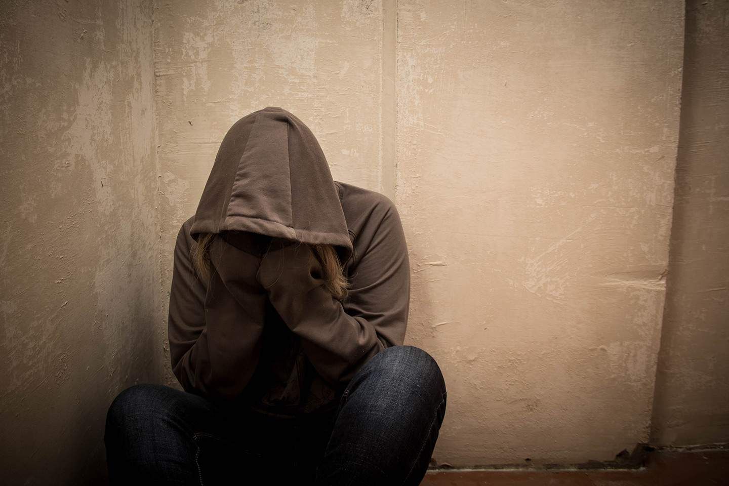 Person with head in hoodie, sitting in corner, hunched over; appears distressed.
