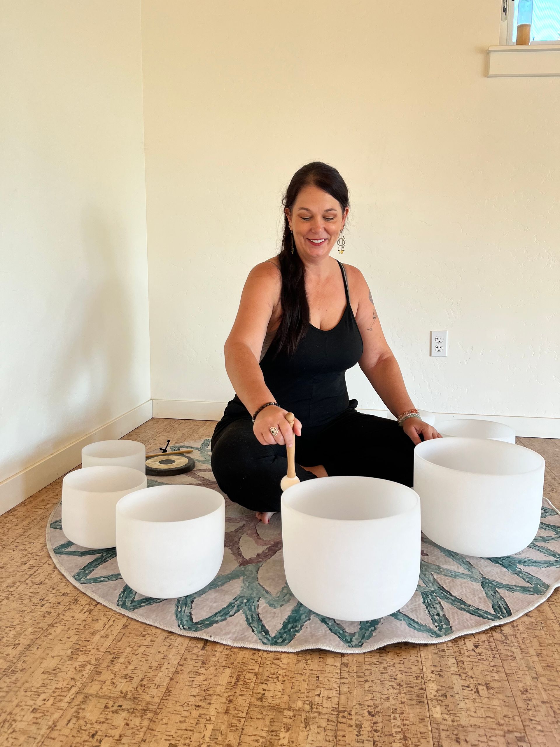 Woman seated, playing crystal singing bowls on a round mat in a light-filled room.