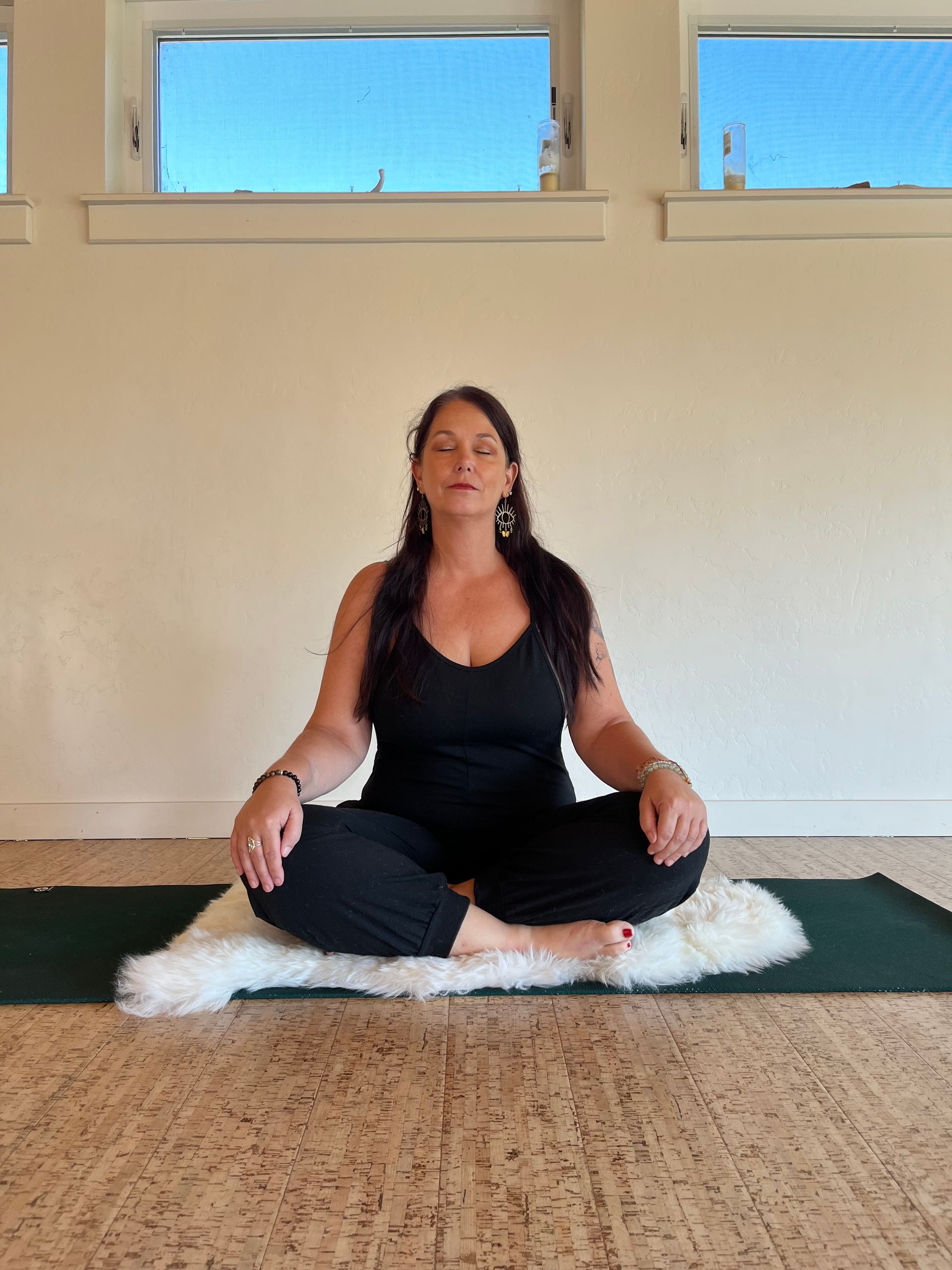 Woman in black clothing meditating indoors, cross-legged on a white sheepskin, eyes closed.