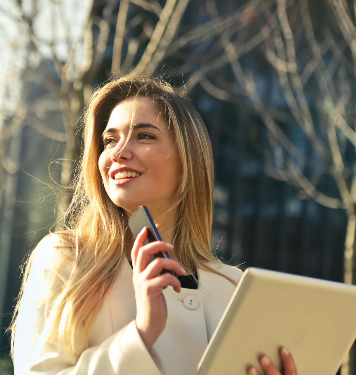 Woman with blonde hair smiles while holding a phone and tablet outdoors.