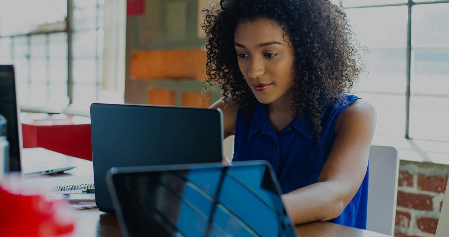 Woman with curly hair in a blue shirt looks at a laptop in an office setting.