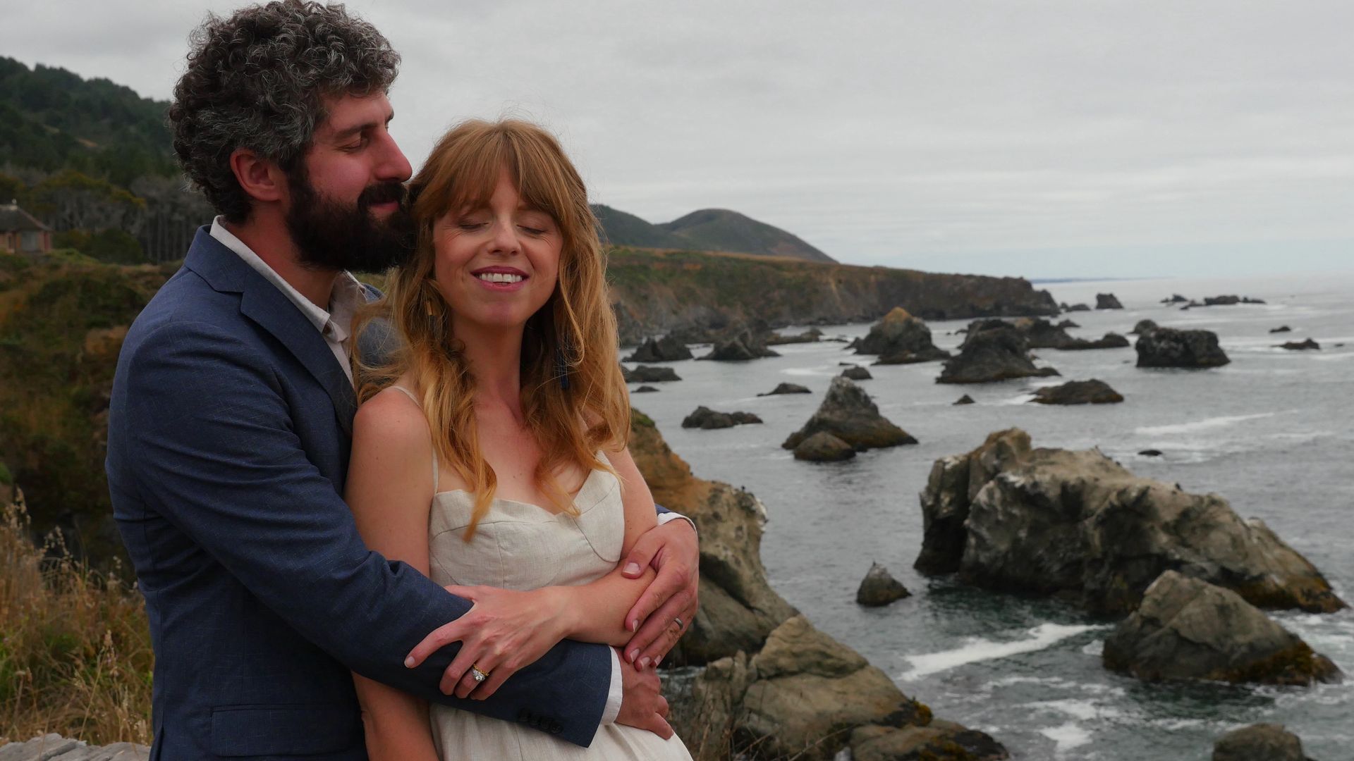 A bride and groom are posing for a picture on a rocky cliff overlooking the ocean.