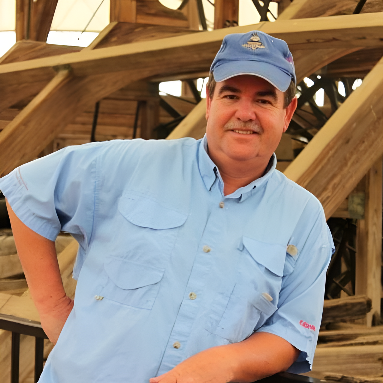 Man in blue shirt and cap, smiling, leaning on railing, wood structure in background.