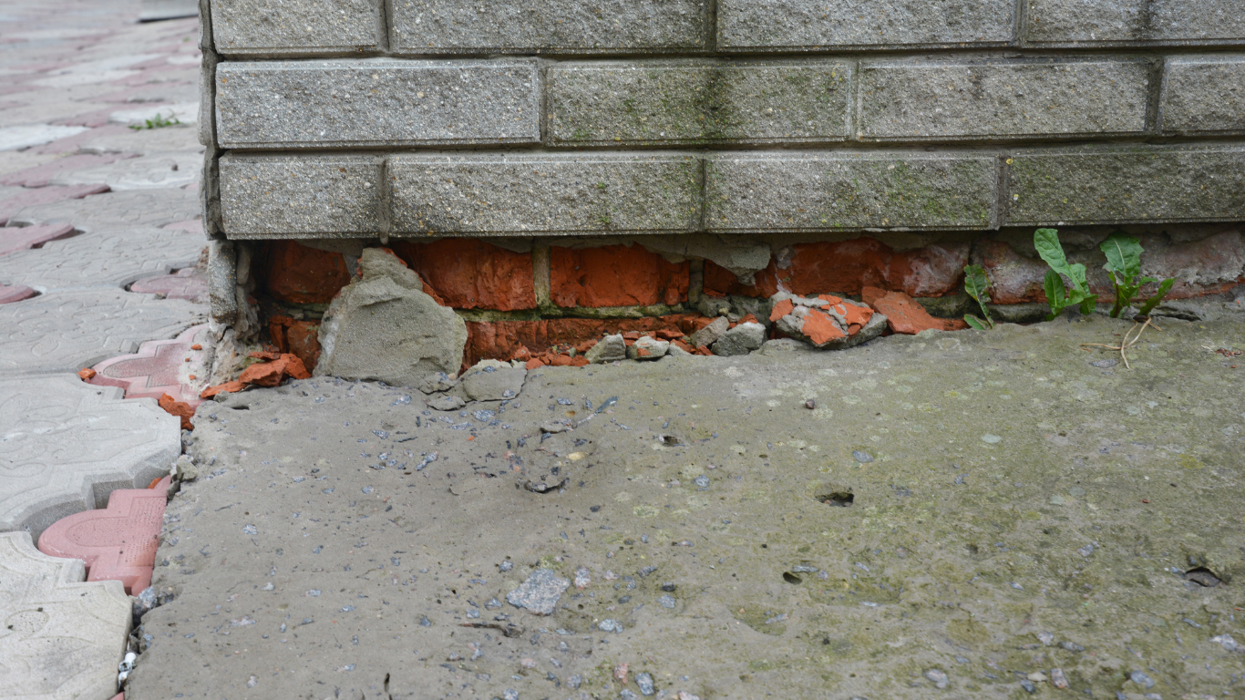 Damaged brick wall base with exposed red bricks, gray concrete, and paving stones.