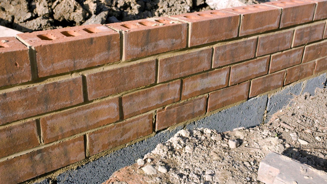 Brick wall construction: red bricks laid with mortar, set on a dark gray base layer, outdoors.