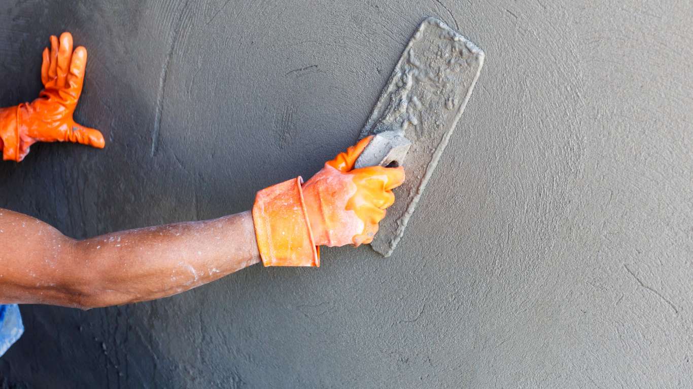 Person wearing orange gloves smoothing cement on a wall with a trowel.