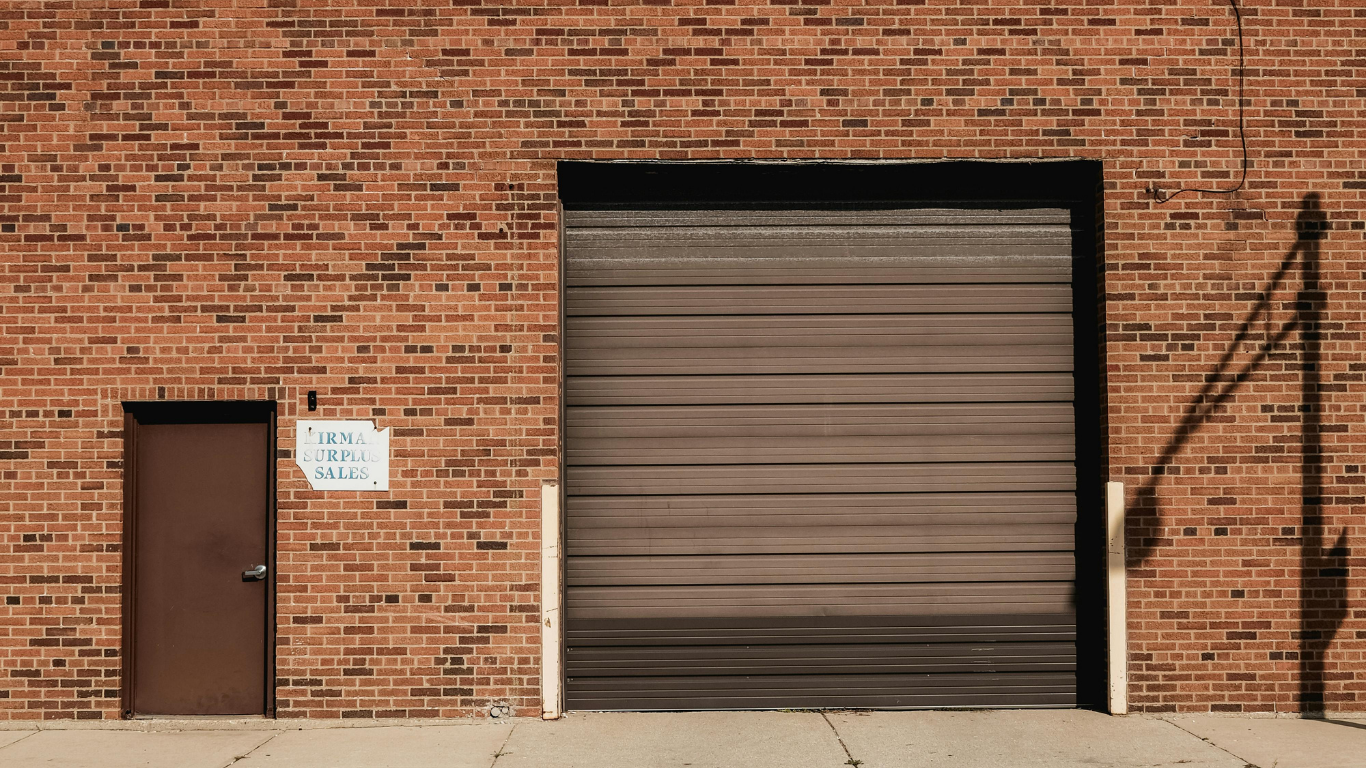 Brick building with brown door, large garage door, and shadow on sidewalk.