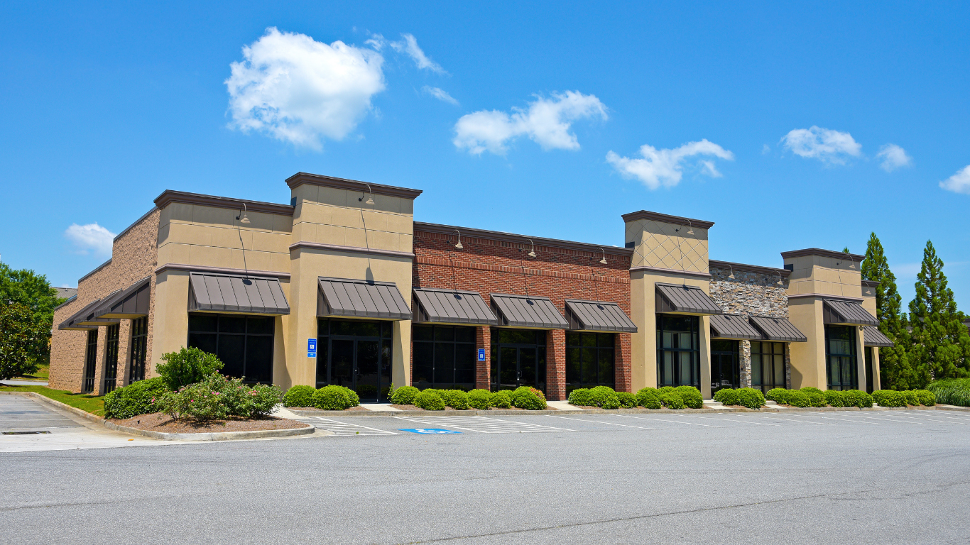 Commercial building with multiple storefronts, awnings, and a paved parking lot under a blue sky.