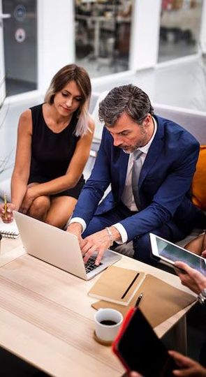 Two business colleagues in a meeting, reviewing a laptop. Man in a suit types; woman watches.