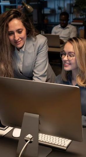 Two women smiling, looking at computer screen in office, another person in the background.