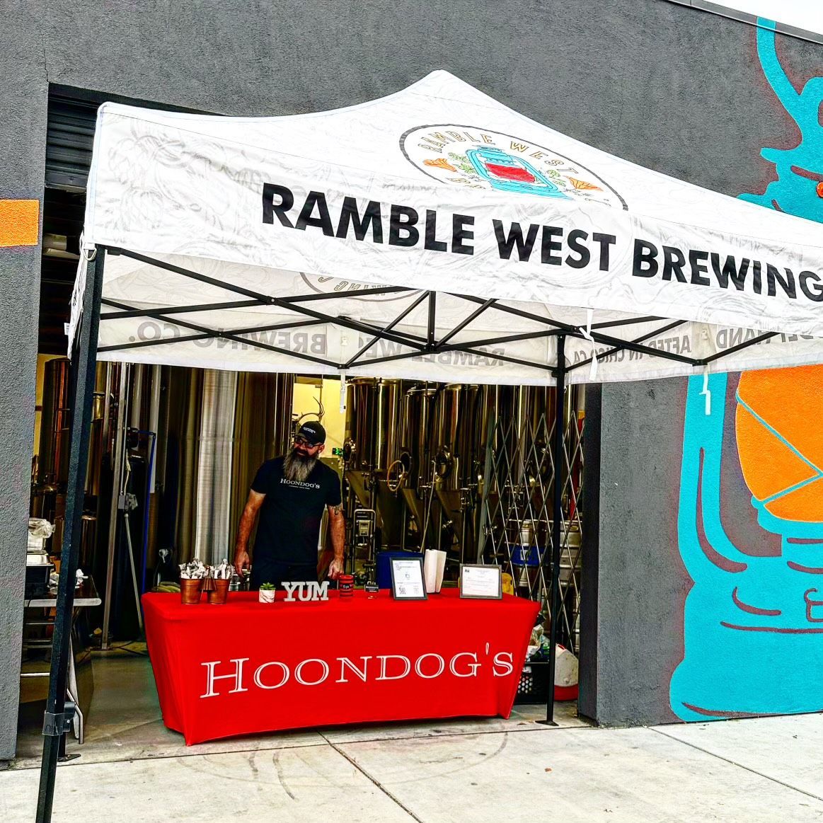a man stands behind a table in front of a ramble west brewing tent