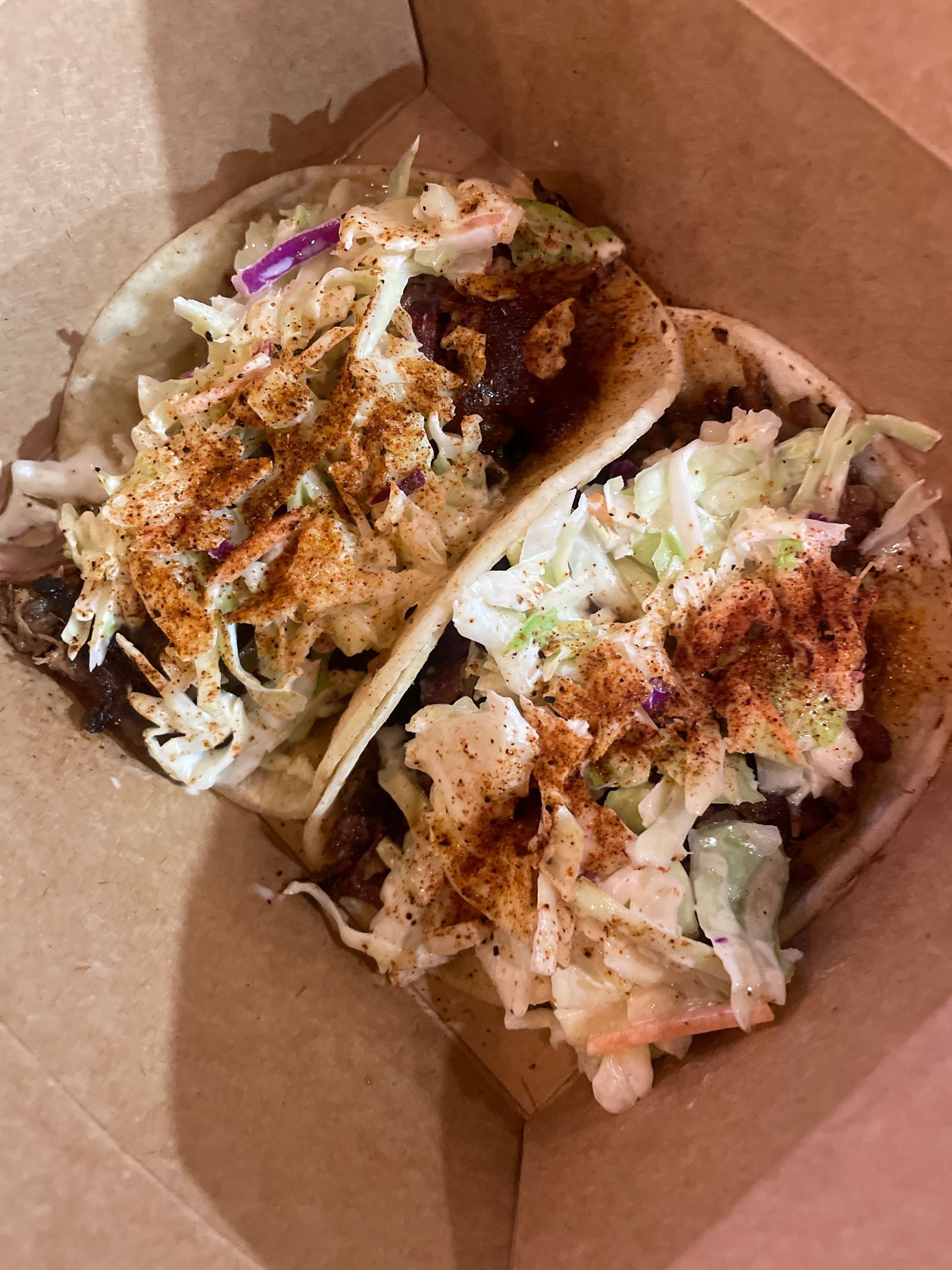 Two tacos in a cardboard container. Each is topped with coleslaw and a reddish spice.