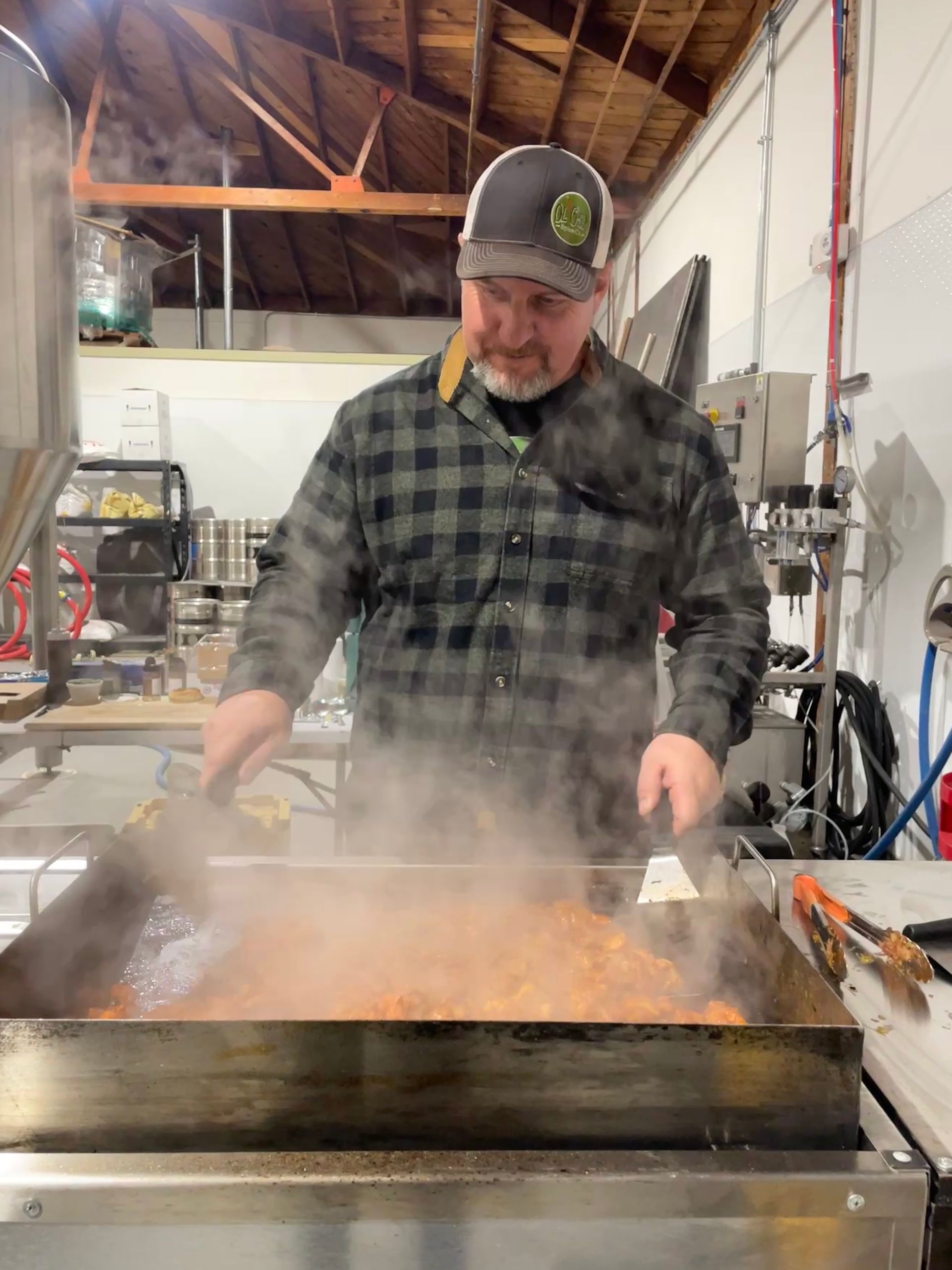 Man in a plaid shirt cooking on a large, steaming flat-top grill inside a commercial kitchen. He is wearing a cap.