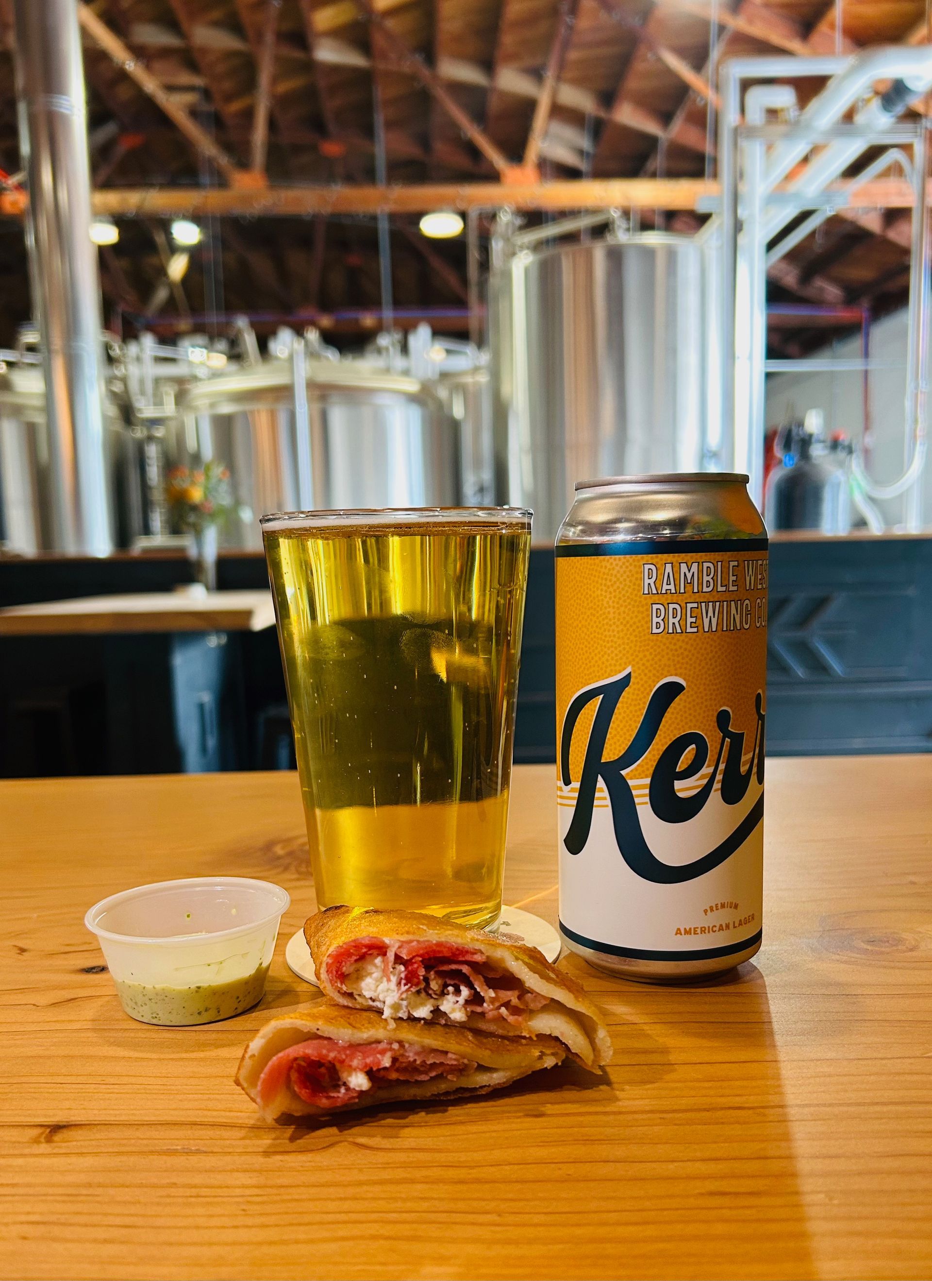 A beer, sandwich, and dipping sauce sit on a wooden table in a brewery. 