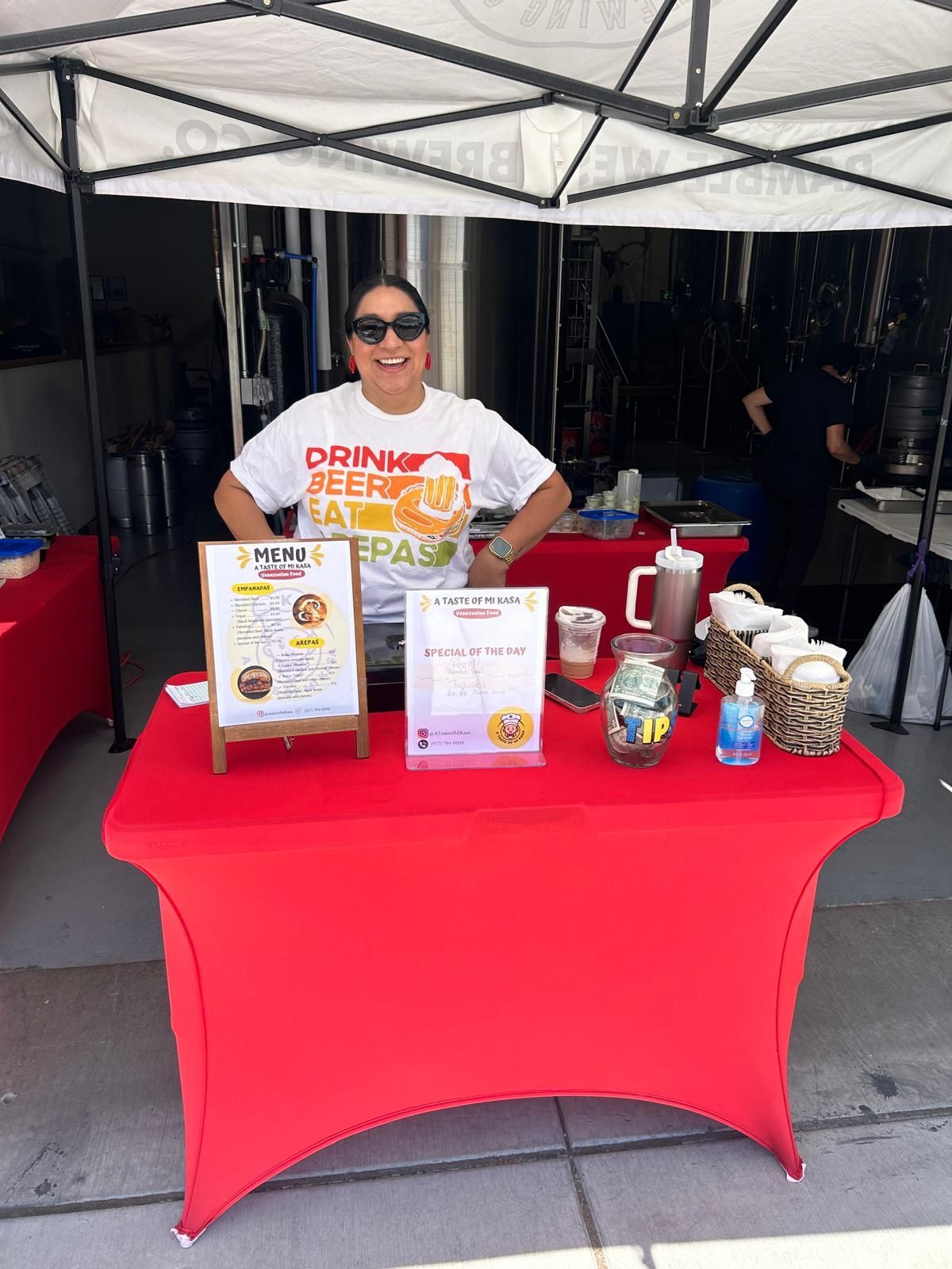Woman smiling at a red-covered table with a menu, drinks, and hand sanitizer. The setting is outdoors under a tent.