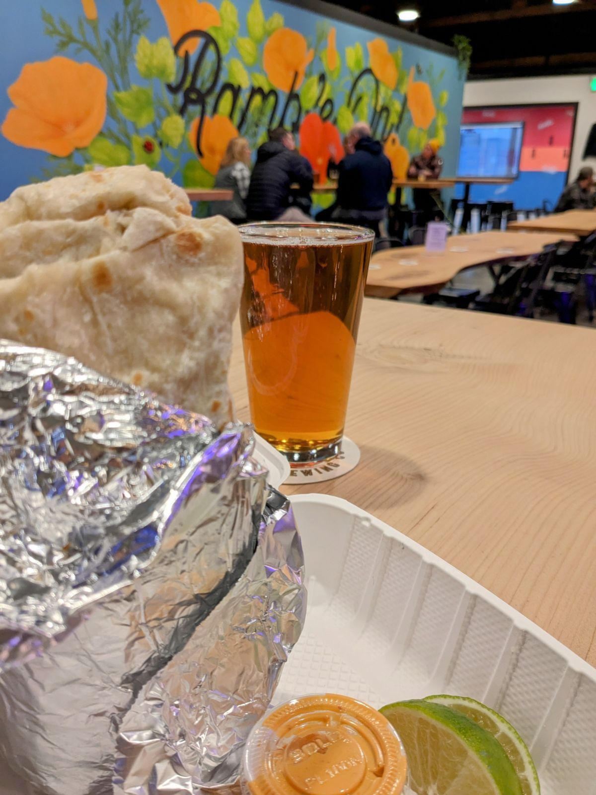 A burrito is wrapped in tin foil next to a glass of beer.