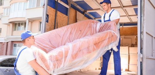 Two movers carry a large, plastic-wrapped couch out of a truck.