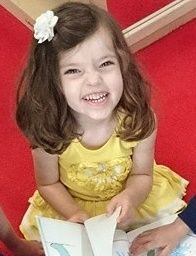 A little girl is sitting on the floor reading a book and smiling.