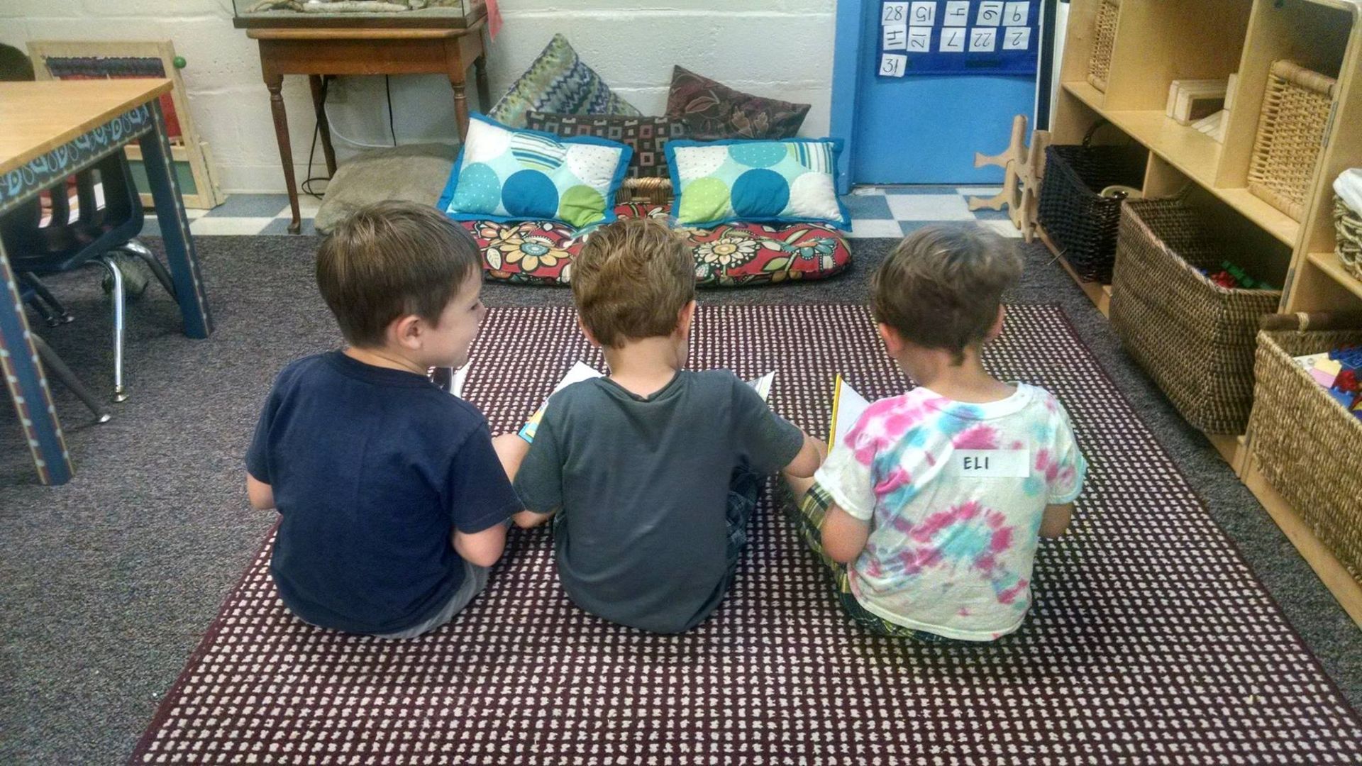 Three children are sitting on the floor reading books in a classroom.