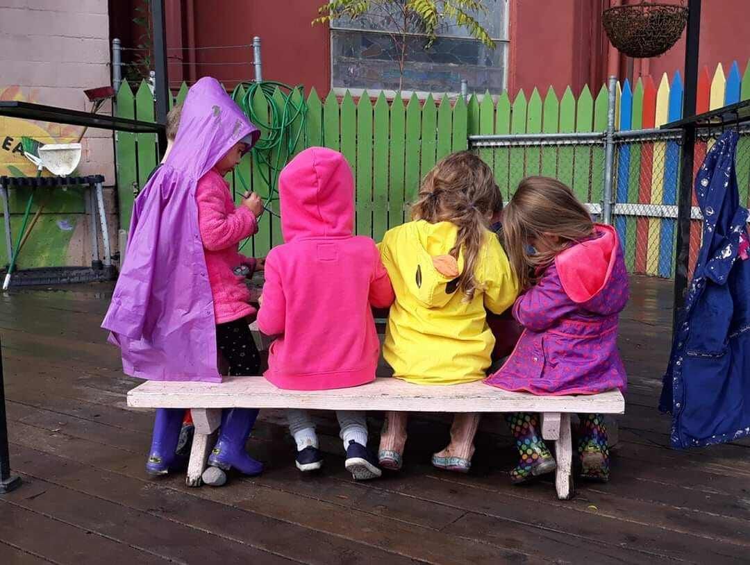 A group of children are sitting on a bench in the rain.