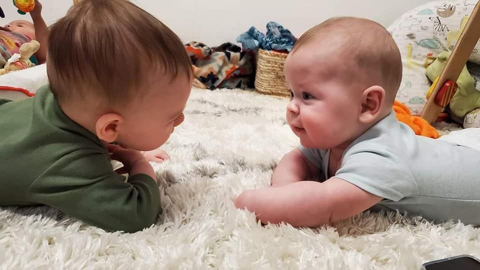 Two babies are laying on their stomachs on a carpet and looking at each other.