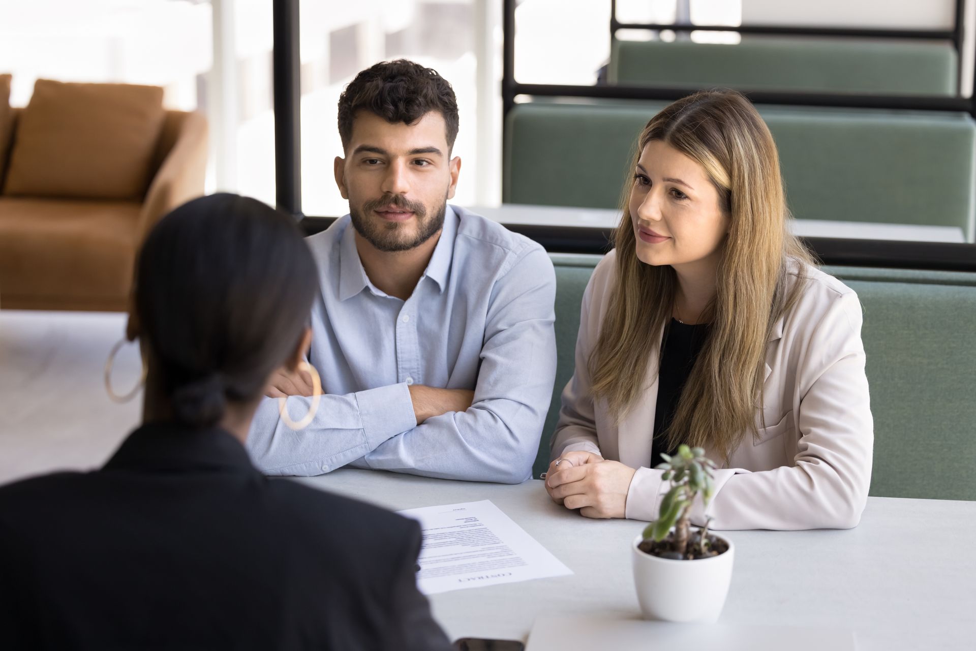 A couple sits across a table from a person with dark hair, possibly in a meeting; indoor, daytime.