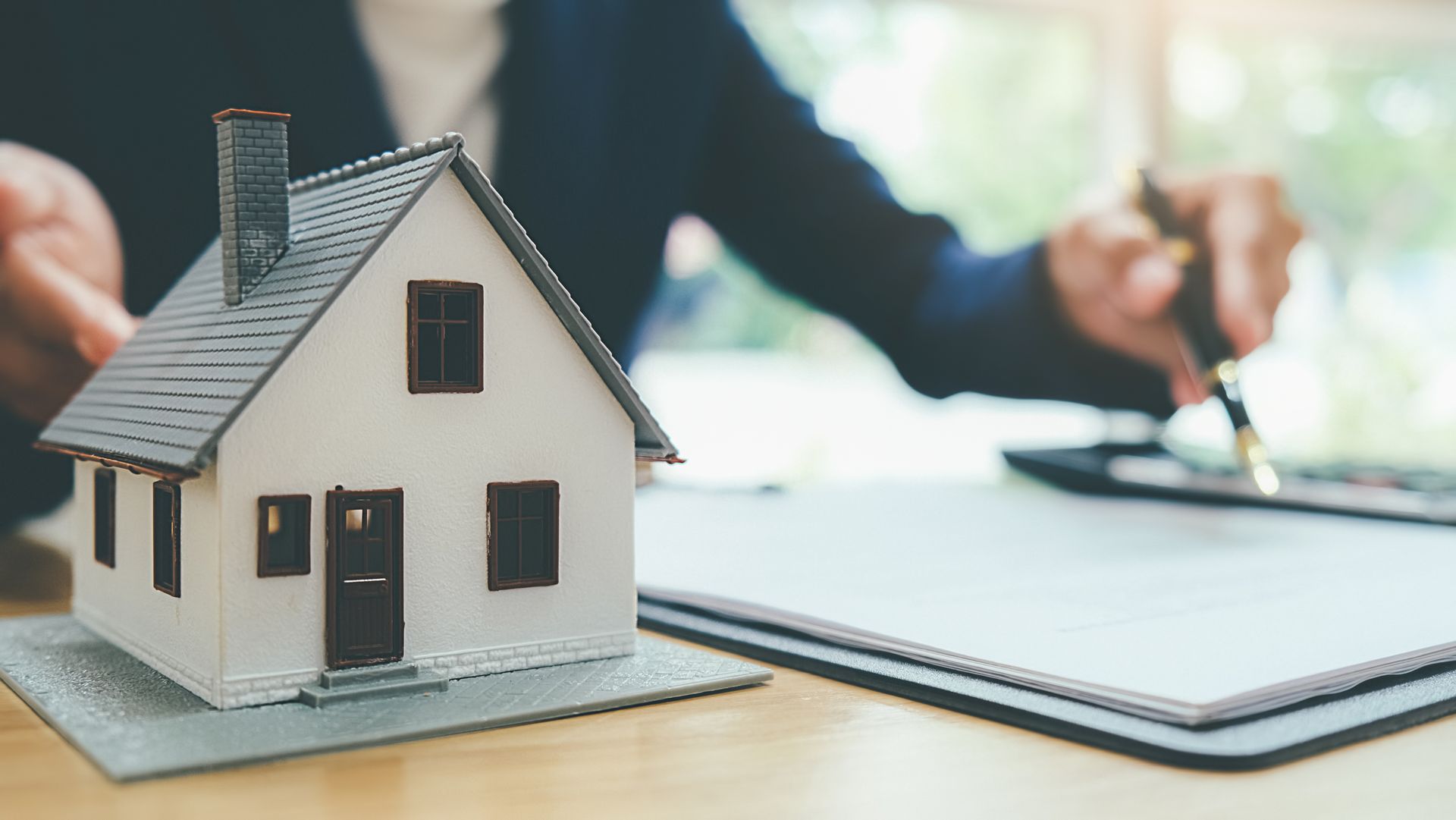 Miniature house on a table next to paperwork and a person's hands holding a pen.