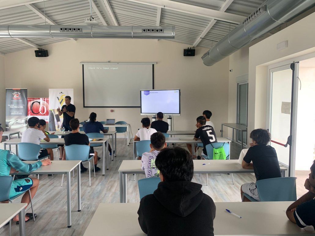 A group of student athletes are sitting at tables in front of a projector screen.