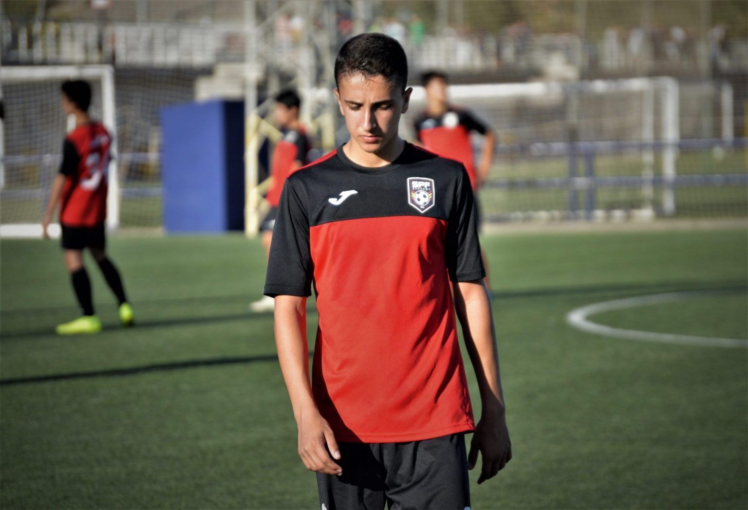 A young man is standing on a soccer field wearing a red and black jersey.