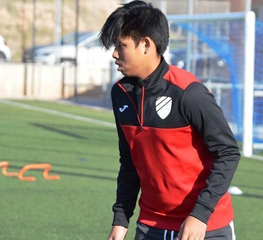 A young man in a red and black jacket stands on a soccer field