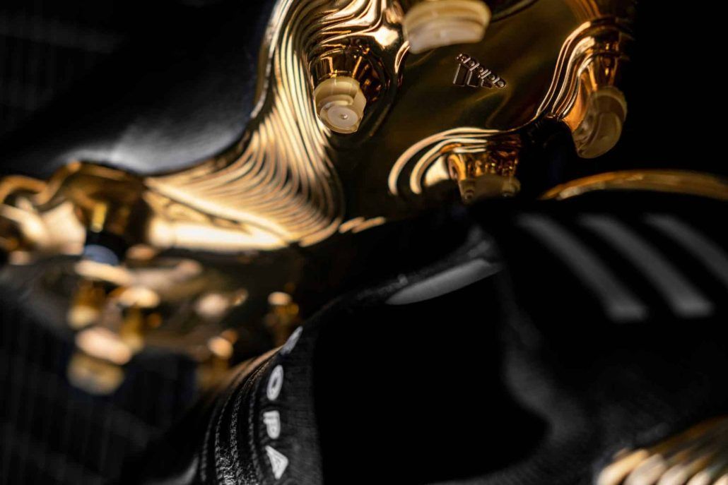 close up image of a black Adidas soccer cleat with a gold bottom and cleats