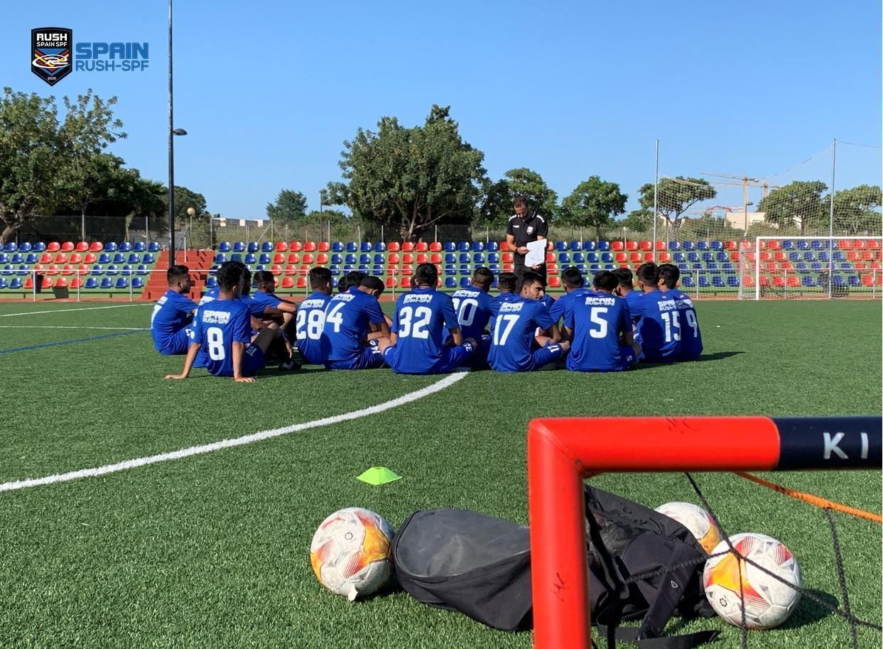 A group of soccer players are sitting on a field with a goal in the foreground.