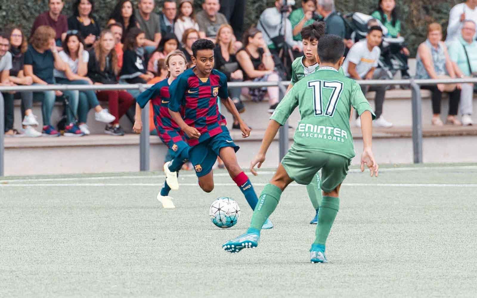 Youth-aged Lamine Yamal dribbles a soccer ball by a player in a green uniform on a soccer field