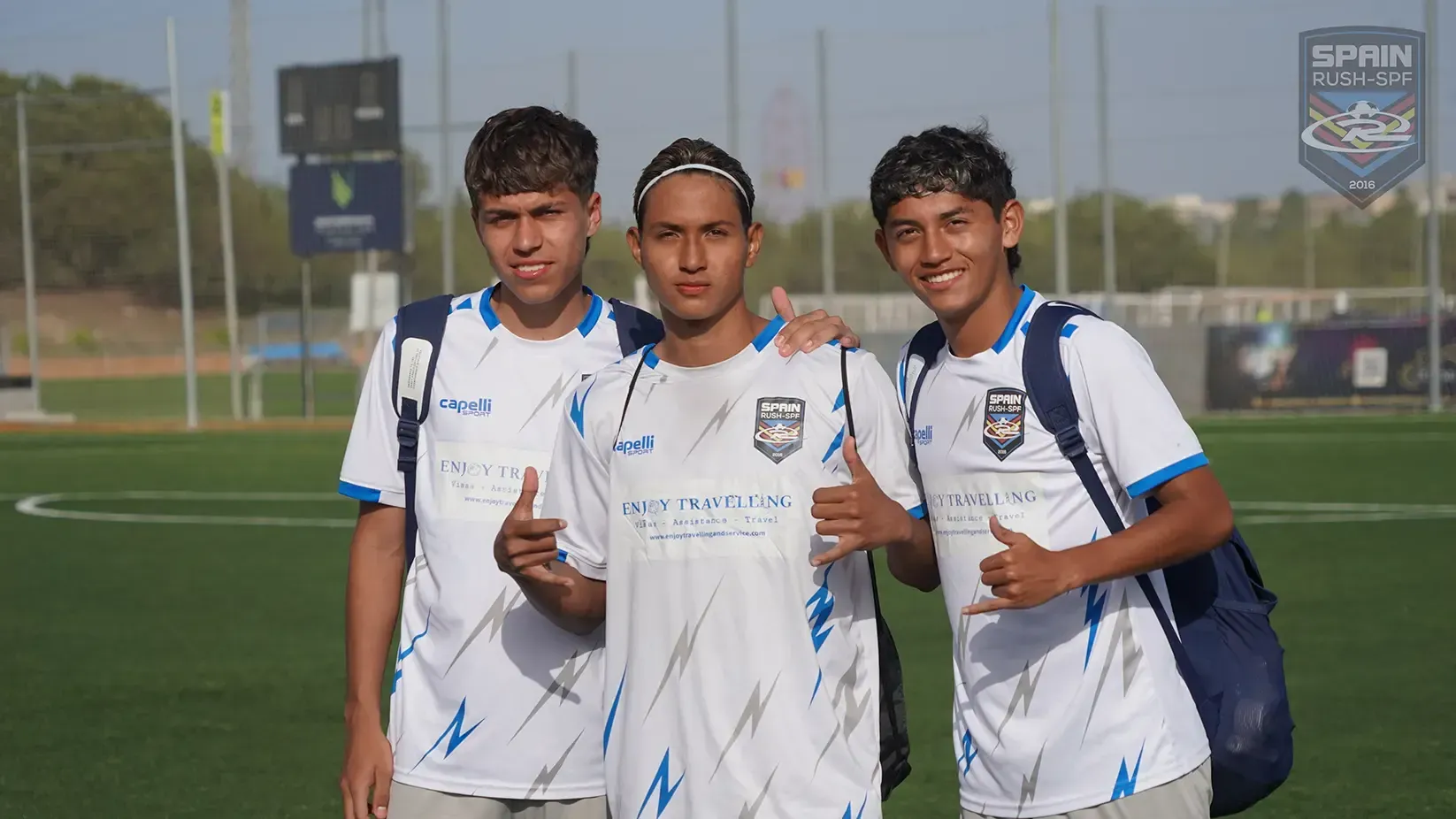 Three soccer players in white jerseys with blue accents, giving thumbs up on a green field.