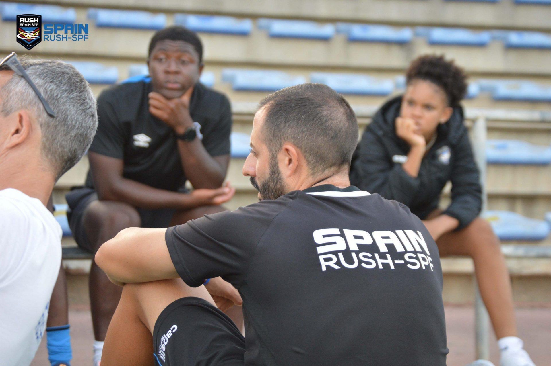 A man wearing a spain rush shirt is sitting on a bench with other people.