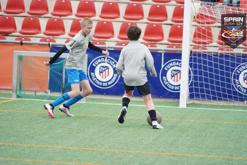 Two young boys are playing soccer on a field with a banner that says spain