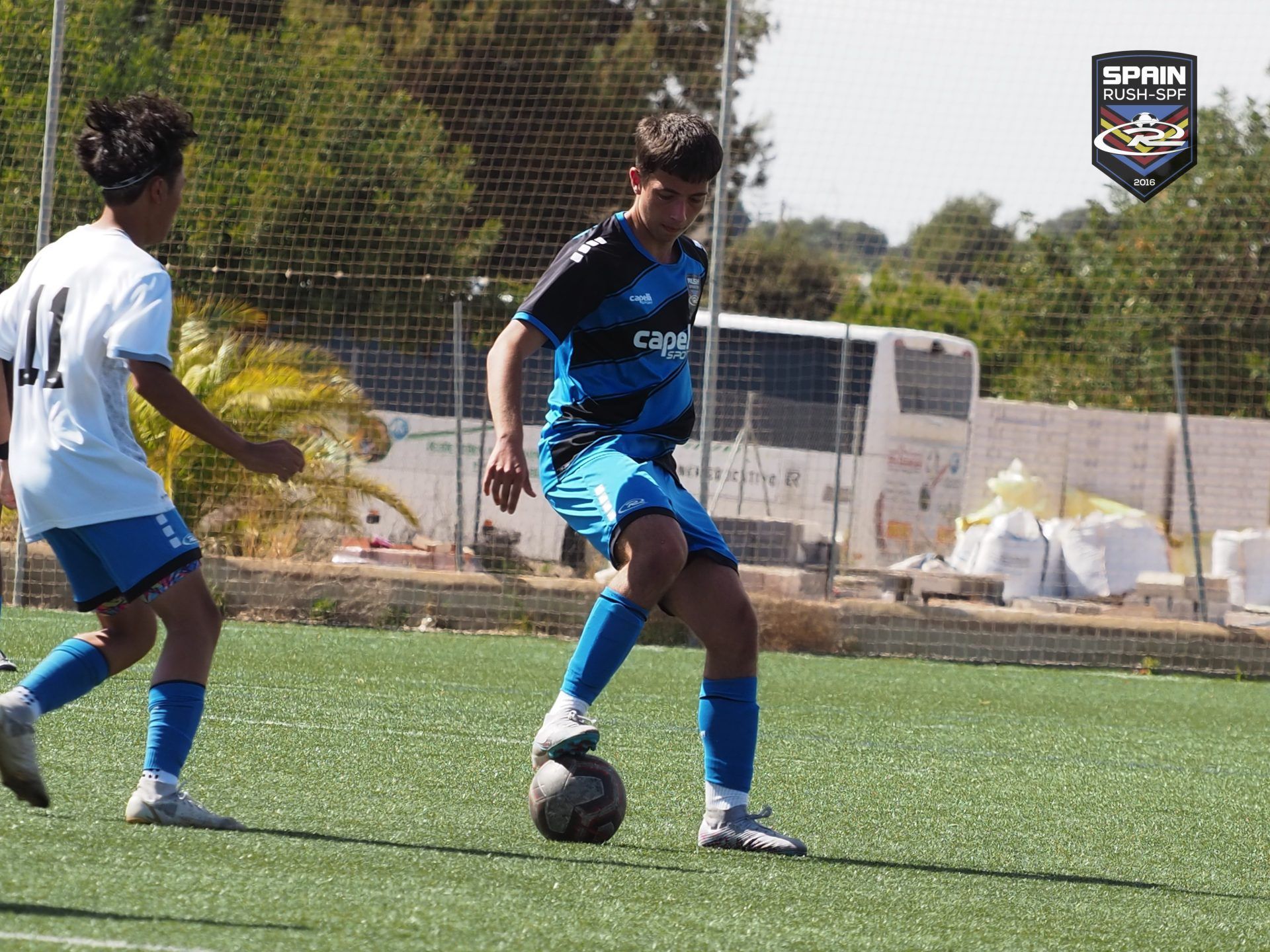 Two boys are playing soccer on a field with a bus in the background.