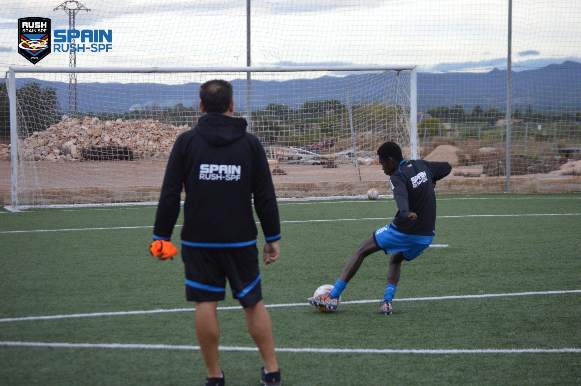 A man is standing on a soccer field watching a man kick a soccer ball.