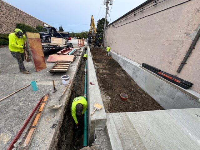 Construction workers installing pipes in a trench alongside a building. One worker is in the trench, another standing nearby, wearing bright safety vests.